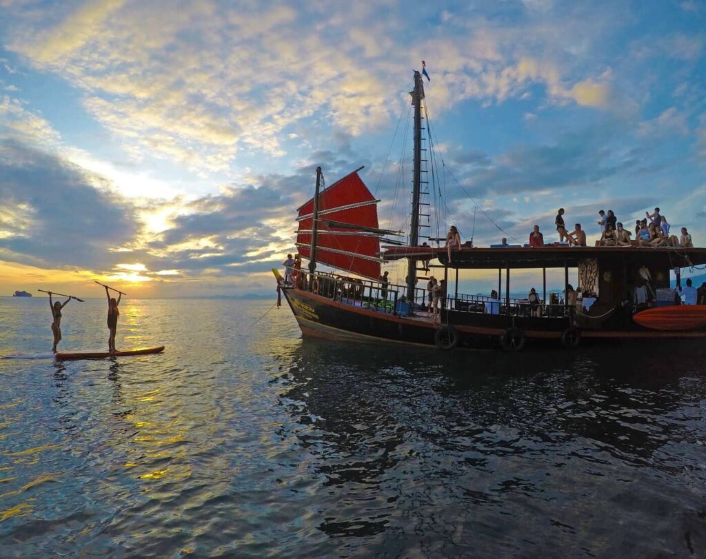 Krabi Sunset Cruises junk boat at sunset with guests on the rooftop and 2 people on paddleboard.