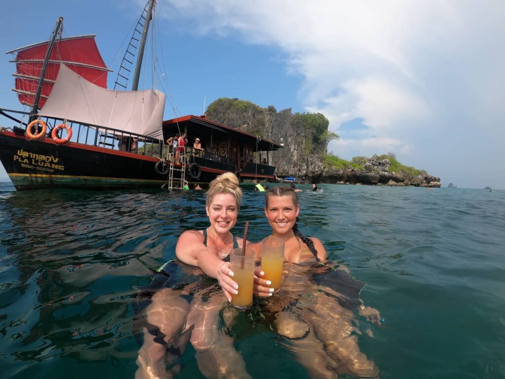 2 girls drinking cocktails in the water with a junk boat behind them.