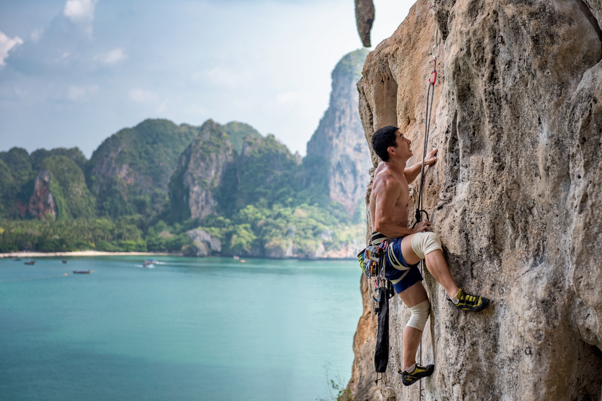 un homme escaladant une paroi rocheuse plate à l'aide de cordes de guidage, avec la plage de Railay et ses falaises de calcaire en arrière-plan à gauche