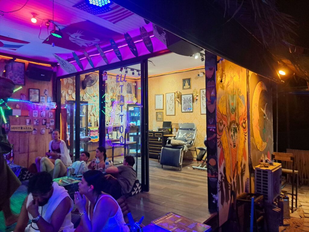 mirando el interior de una tienda de tatuajes en railay, krabi. la puerta está abierta y hay clientes del bar sentados fuera.