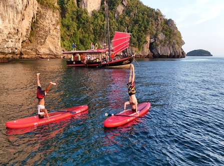 Two guests performing handstands on red paddleboards with the red-sailed junk boat and limestone cliffs in the background