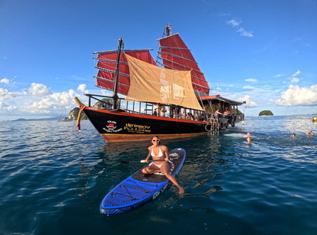 Guest on a paddleboard beside the red-sailed junk boat with others swimming nearby