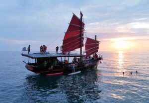Un bateau de « Krabi Sunset Cruises » au coucher du soleil, avec des clients faisant du paddle à ses côtés et d'autres admirant le coucher de soleil depuis le toit