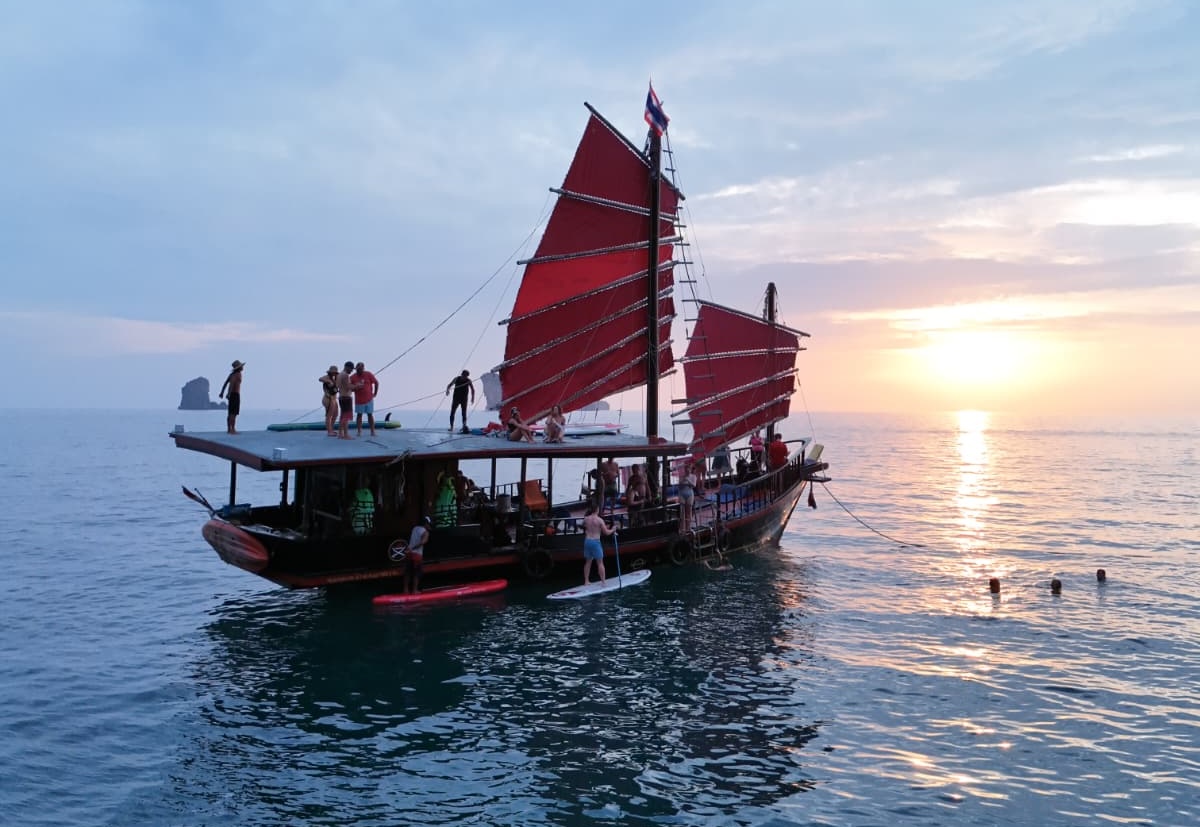 Un bateau de « Krabi Sunset Cruises » au coucher du soleil, avec des clients faisant du paddle à ses côtés et d'autres admirant le coucher de soleil depuis le toit